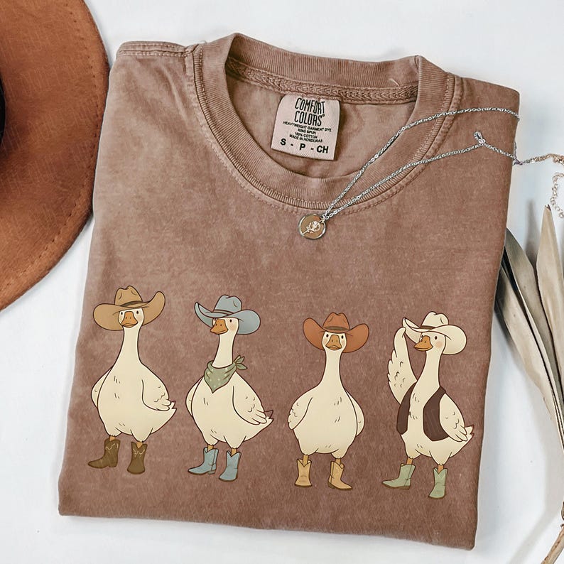 May include: A brown Comfort Colors t-shirt featuring a graphic of four geese wearing cowboy hats and boots. The geese are in various poses, with one wearing a vest and another a bandana. A silver necklace is draped across the shirt.