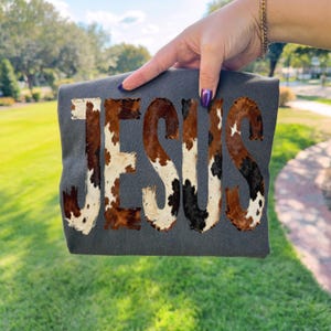 K&ouml;nnte beinhalten: Ein dunkelgraues T-Shirt mit dem Wort "JESUS" in einem Kuhfellmuster. Die Buchstaben sind eine Mischung aus Braun, Schwarz und Wei&szlig; und erzeugen ein rustikales Design. Das T-Shirt wird vor einem gr&uuml;nen, grasbewachsenen Hintergrund gehalten.