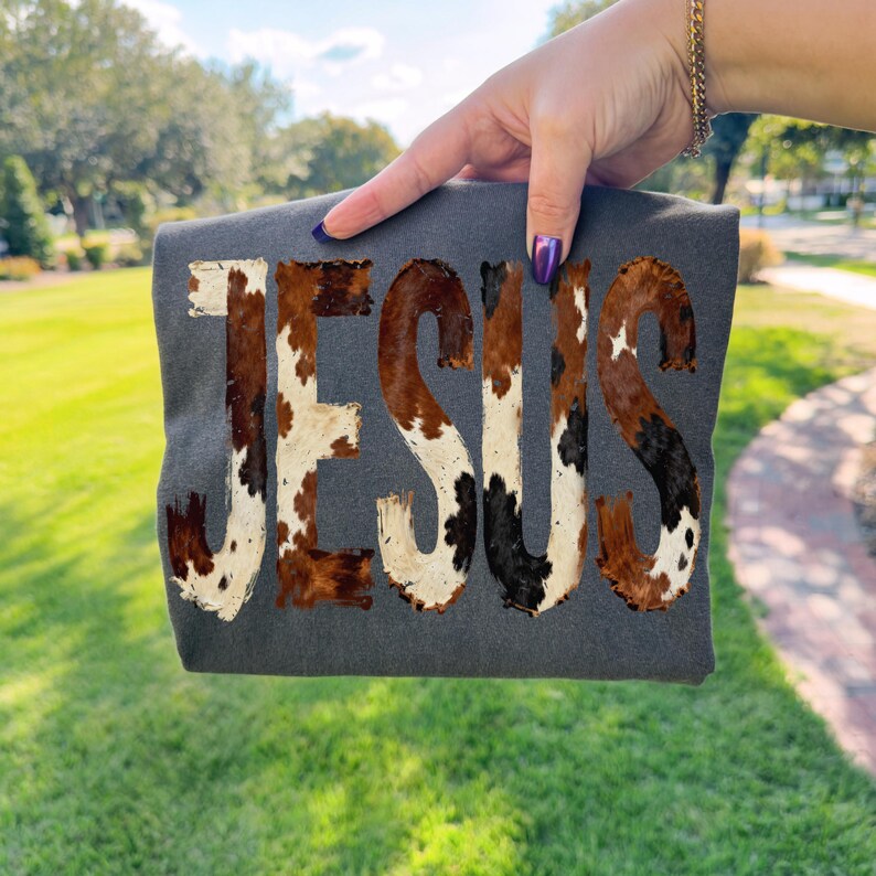K&ouml;nnte beinhalten: Ein dunkelgraues T-Shirt mit dem Wort "JESUS" in einem Kuhfellmuster. Die Buchstaben sind eine Mischung aus Braun, Schwarz und Wei&szlig; und erzeugen ein rustikales Design. Das T-Shirt wird vor einem gr&uuml;nen, grasbewachsenen Hintergrund gehalten.