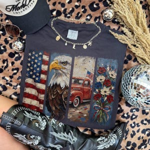 Puede incluir: Camiseta gris oscuro con un dise&ntilde;o patri&oacute;tico que presenta la bandera estadounidense, un &aacute;guila, una camioneta roja y flores. La camiseta est&aacute; adornada con un collar dorado, gafas de sol y una gorra negra. Tambi&eacute;n se ven botas de vaquero en blanco y negro.