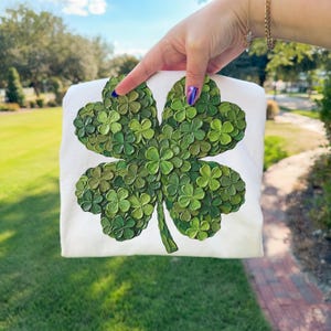 May include: White sweatshirt featuring a large, textured four-leaf clover design in shades of green. The clover is composed of many small, three-dimensional shamrocks. The background is a green lawn and a blue sky.