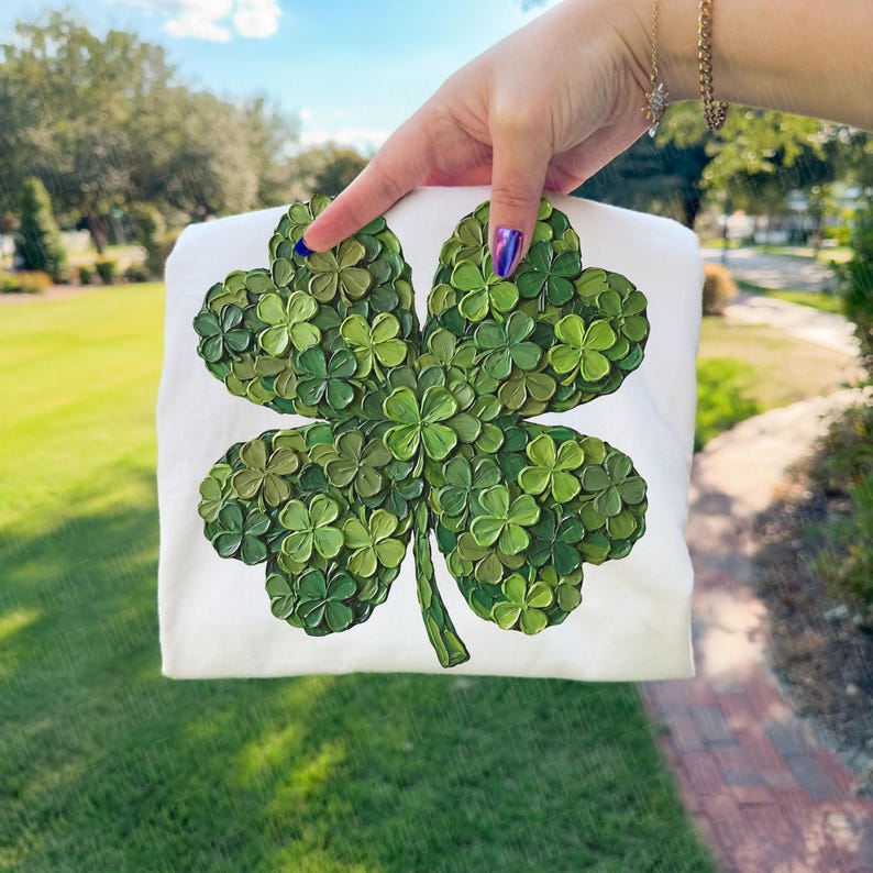 May include: White sweatshirt featuring a large, textured four-leaf clover design in shades of green. The clover is composed of many small, three-dimensional shamrocks. The background is a green lawn and a blue sky.