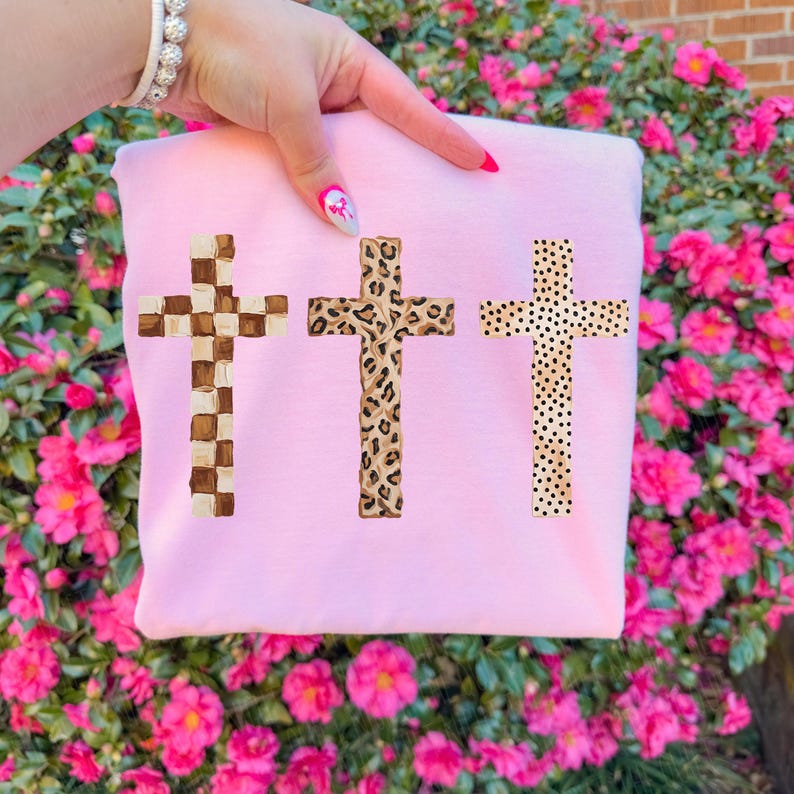 May include: A light pink sweatshirt featuring three crosses with unique patterns. The crosses include a checkered design, a leopard print, and a dotted pattern. The sweatshirt is held up by a person's hand.