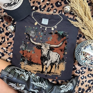 May include: A dark gray t-shirt featuring a longhorn steer illustration with a desert landscape background. The image is surrounded by a black cap, sunglasses, a gold necklace, a pair of black cowboy boots, and disco balls.