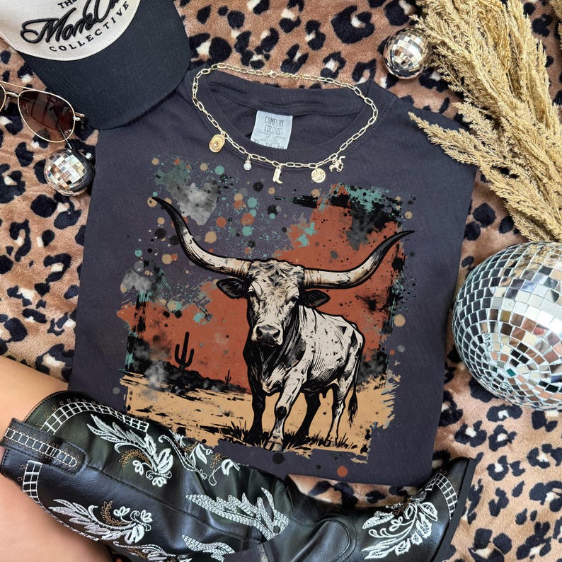 May include: A dark gray t-shirt featuring a longhorn steer illustration with a desert landscape background. The image is surrounded by a black cap, sunglasses, a gold necklace, a pair of black cowboy boots, and disco balls.