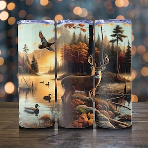 May include: Three stainless steel insulated tumblers with a nature scene. The design features a deer, ducks, and a lake surrounded by trees. The color palette includes shades of brown, orange, and green, with a touch of blue.