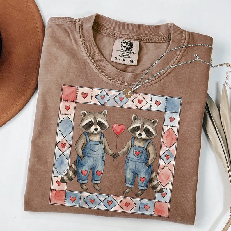 Pu&ograve; includere: T-shirt Comfort Colors marrone con una grafica ad acquerello di due procioni che si tengono per mano e un palloncino a forma di cuore rosso. I procioni indossano salopette blu. Una collana d'argento con un ciondolo &egrave; drappeggiata sulla maglietta.