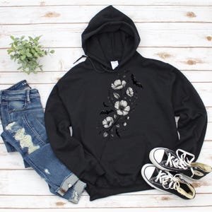 May include: Black hoodie with a floral and bat design. The hoodie features a drawstring hood and a front pocket. The floral design is in grayscale, with white flowers and black bats. A pair of black and white sneakers and blue jeans are also in the image.