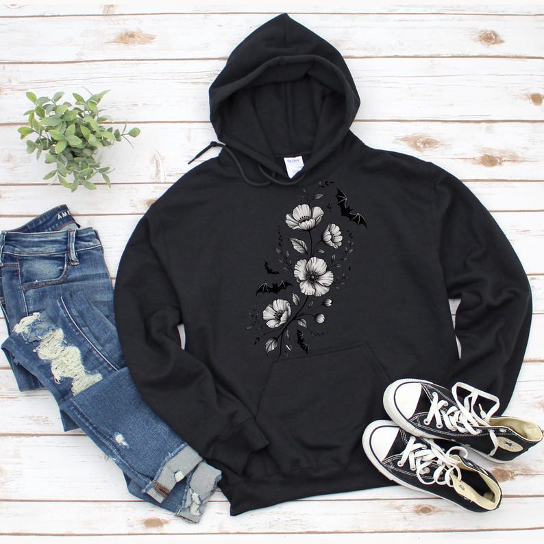 May include: Black hoodie with a floral and bat design. The hoodie features a drawstring hood and a front pocket. The floral design is in grayscale, with white flowers and black bats. A pair of black and white sneakers and blue jeans are also in the image.