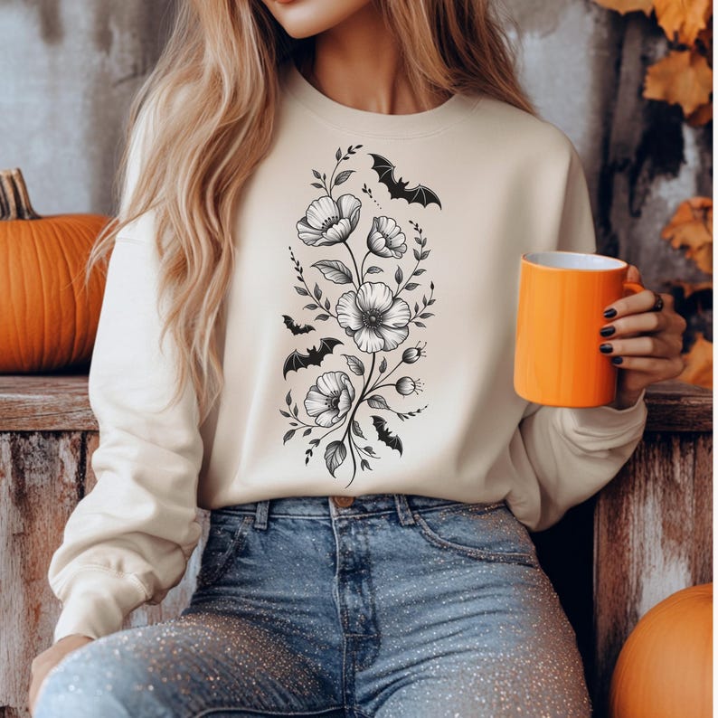 May include: Cream-colored sweatshirt featuring a black floral and bat design. The graphic includes detailed flowers and bats, creating a Halloween or gothic aesthetic. The person is holding an orange mug.