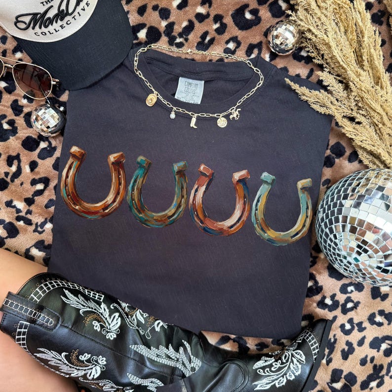 May include: Black t-shirt with a horseshoe design in shades of brown, blue, and red. The shirt is accessorized with a gold necklace, a black cap, sunglasses, and a pair of black cowboy boots. A disco ball and decorative wheat are also present.
