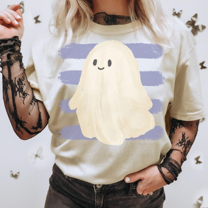 May include: A cream-colored short-sleeve t-shirt featuring a central graphic of a pale yellow ghost with a simple black-lined face. The ghost is positioned over a background of four horizontal, distressed-texture light purple brush strokes.