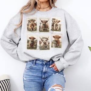 May include: A light gray sweatshirt featuring a collage of six Highland cow illustrations. Each cow is depicted in a vintage postcard style, with details like floral crowns and rustic farm settings. The sweatshirt is made of a soft material.