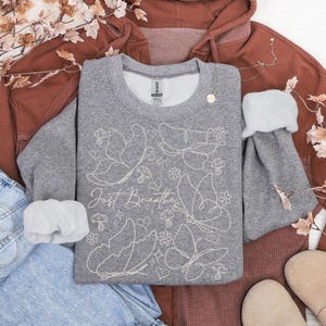 Puede incluir: Una sudadera gris con un dise&ntilde;o de mariposas y flores blancas, y la frase "Just Breathe". La sudadera est&aacute; rodeada por una sudadera con capucha de color &oacute;xido, unos vaqueros azules y unos zapatos beige. Tambi&eacute;n hay un collar dorado y hojas decorativas.