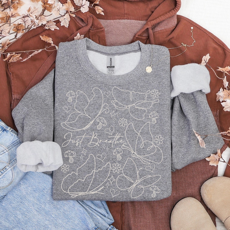 Puede incluir: Una sudadera gris con un dise&ntilde;o de mariposas y flores blancas, y la frase "Just Breathe". La sudadera est&aacute; rodeada por una sudadera con capucha de color &oacute;xido, unos vaqueros azules y unos zapatos beige. Tambi&eacute;n hay un collar dorado y hojas decorativas.