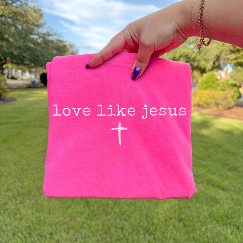 May include: A bright pink t-shirt with the words "love like jesus" and a white cross printed on the front. The shirt is held up against a background of green grass and trees.