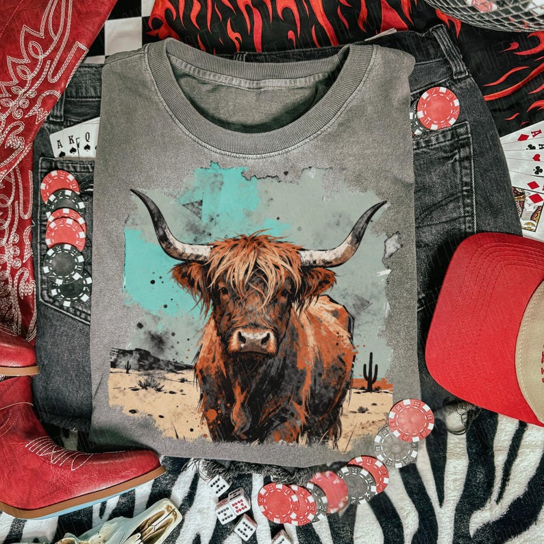 May include: A grey t-shirt with a watercolour illustration of a Highland cow. The image is surrounded by denim jeans, a red cap, red boots, playing cards, and poker chips, suggesting a Western theme.