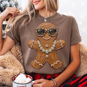 May include: A brown short-sleeve t-shirt featuring a graphic of a gingerbread person wearing leopard print sunglasses and a silver-toned chain necklace. The gingerbread figure has a textured, impasto-style paint appearance with white icing details.