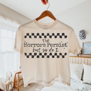 May include: Cream-colored t-shirt with the text "The Horrors Persist but so do I" in black, framed by a black and white checkered pattern. The shirt is hanging on a wooden hanger.