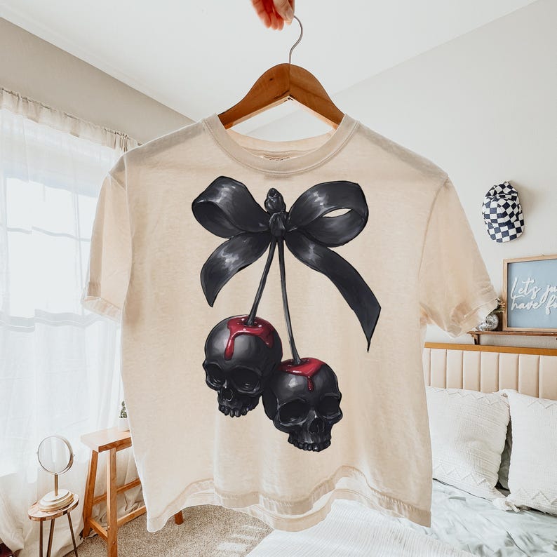 May include: A cream-coloured t-shirt with a graphic of two black skull-shaped cherries with red drips, connected by stems to a large black bow. The shirt is hanging on a wooden hanger.