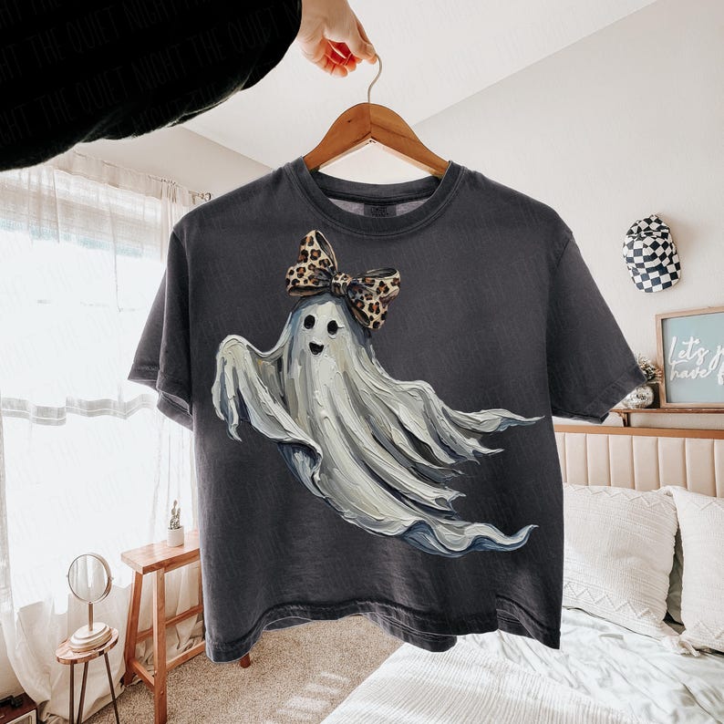 May include: A dark grey t-shirt with a whimsical ghost design. The ghost has a light grey body and a smiling face, decorated with a leopard print bow. The t-shirt is hanging on a wooden hanger.