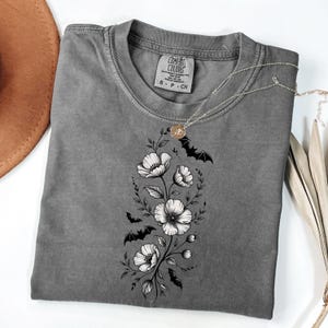 May include: A gray Comfort Colors t-shirt with a floral and bat design. The graphic features white flowers and black bats arranged vertically. A silver necklace with a pendant is draped over the shirt.