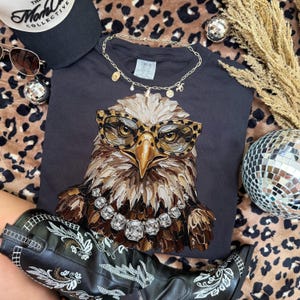 May include: A black t-shirt featuring a detailed illustration of an eagle wearing leopard print glasses and a diamond necklace. The image includes a black cap, sunglasses, a gold necklace, and black cowboy boots with white embroidery.