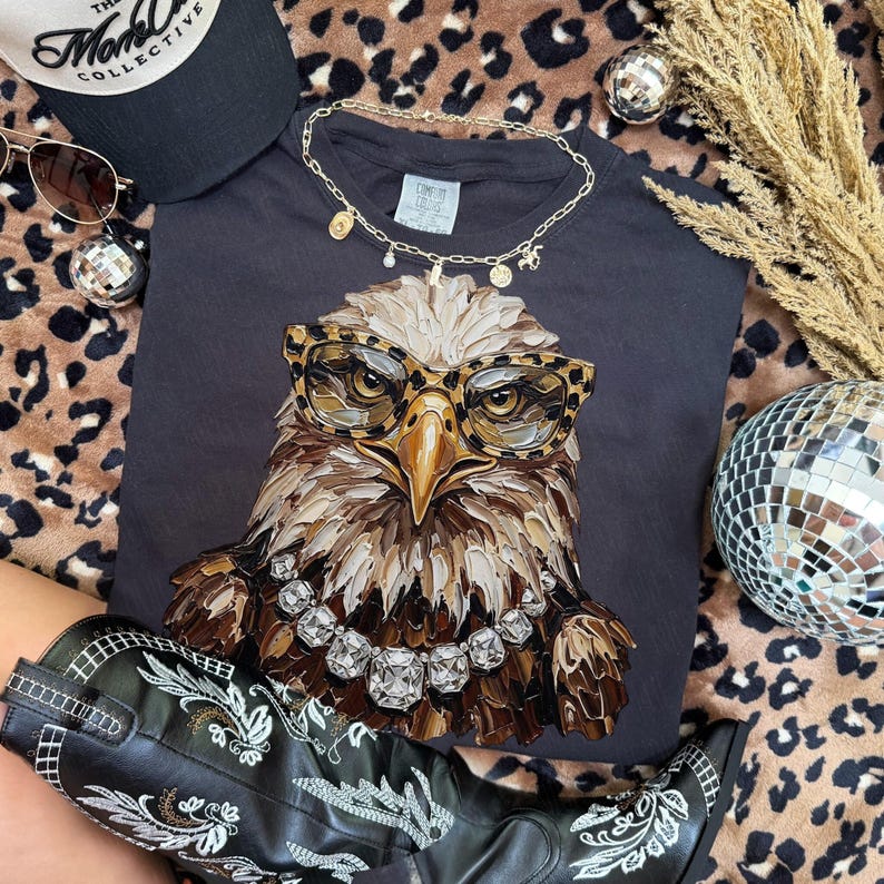 May include: A black t-shirt featuring a detailed illustration of an eagle wearing leopard print glasses and a diamond necklace. The image includes a black cap, sunglasses, a gold necklace, and black cowboy boots with white embroidery.