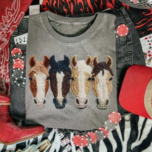 Peut inclure: Un sweat-shirt gris &agrave; col rond avec un motif brod&eacute; de quatre t&ecirc;tes de chevaux dans les tons marron, noir et blanc. Le sweat-shirt est entour&eacute; d'un jean, d'un chapeau de cowboy rouge et de cartes &agrave; jouer, sugg&eacute;rant un th&egrave;me western.