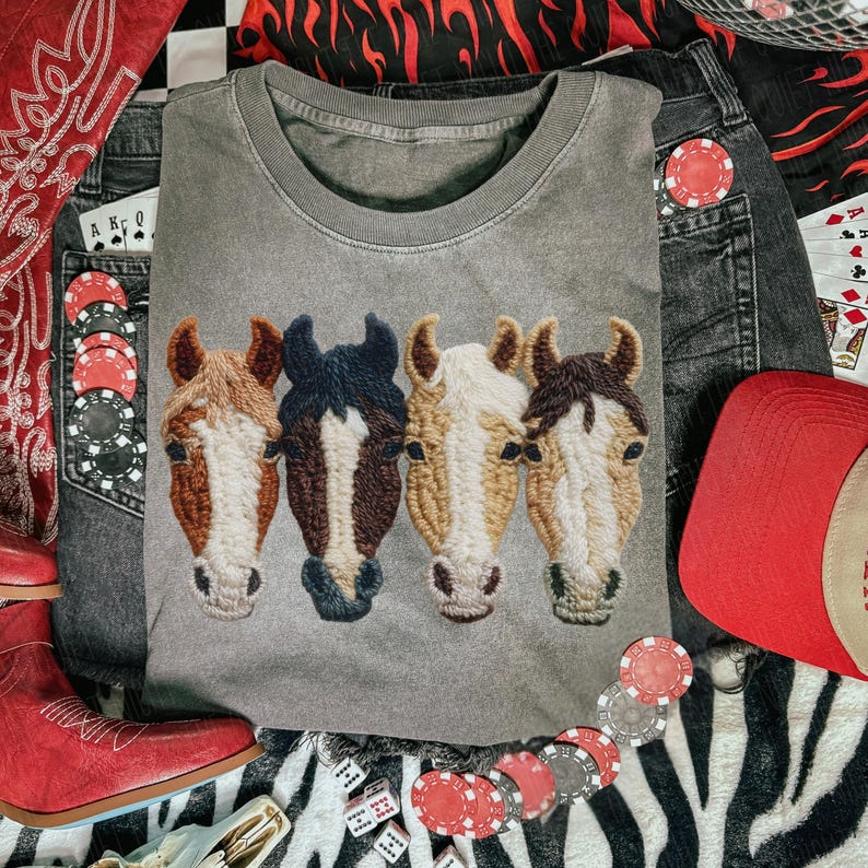 Peut inclure: Un sweat-shirt gris &agrave; col rond avec un motif brod&eacute; de quatre t&ecirc;tes de chevaux dans les tons marron, noir et blanc. Le sweat-shirt est entour&eacute; d'un jean, d'un chapeau de cowboy rouge et de cartes &agrave; jouer, sugg&eacute;rant un th&egrave;me western.