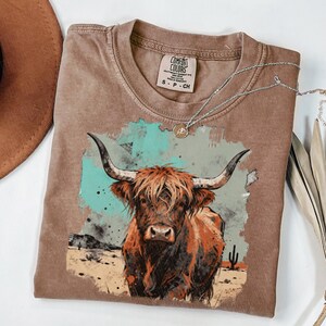 May include: A brown Comfort Colors t-shirt with a Highland cow graphic, set against a desert scene with a turquoise and grey watercolour background. A silver necklace with a pendant is draped on the shirt. The label reads S-P-CH.