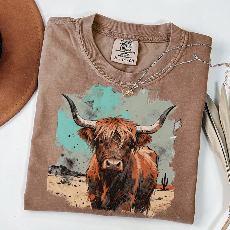 May include: A brown Comfort Colors t-shirt with a Highland cow graphic, set against a desert scene with a turquoise and grey watercolour background. A silver necklace with a pendant is draped on the shirt. The label reads S-P-CH.