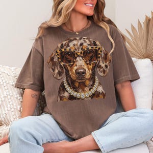 May include: A brown short-sleeve t-shirt featuring a graphic of a dapple dachshund wearing leopard print glasses and a chunky gold chain necklace with clear stones. The dog has a mottled brown and tan coat and is centered on the chest.