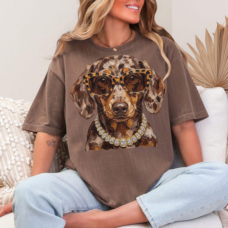 May include: A brown short-sleeve t-shirt featuring a graphic of a dapple dachshund wearing leopard print glasses and a chunky gold chain necklace with clear stones. The dog has a mottled brown and tan coat and is centered on the chest.