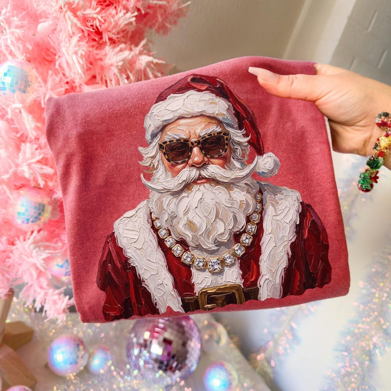 May include: A dusty rose-colored t-shirt featuring a graphic print of Santa Claus wearing leopard print sunglasses and a chunky gold chain necklace. The artwork has a textured, impasto-style paint appearance with visible brushstrokes.
