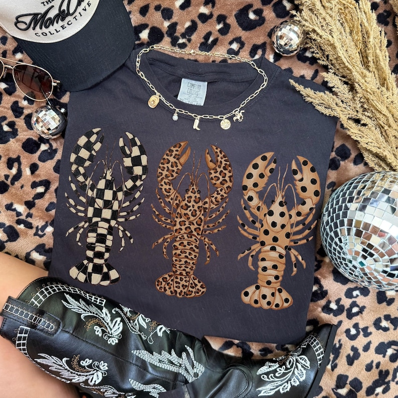 May include: Dark gray t-shirt featuring three lobster designs with different patterns: checkered, leopard print, and spotted. The shirt is accessorized with a gold necklace, sunglasses, and a black cap. A pair of black and white embroidered boots are also visible.