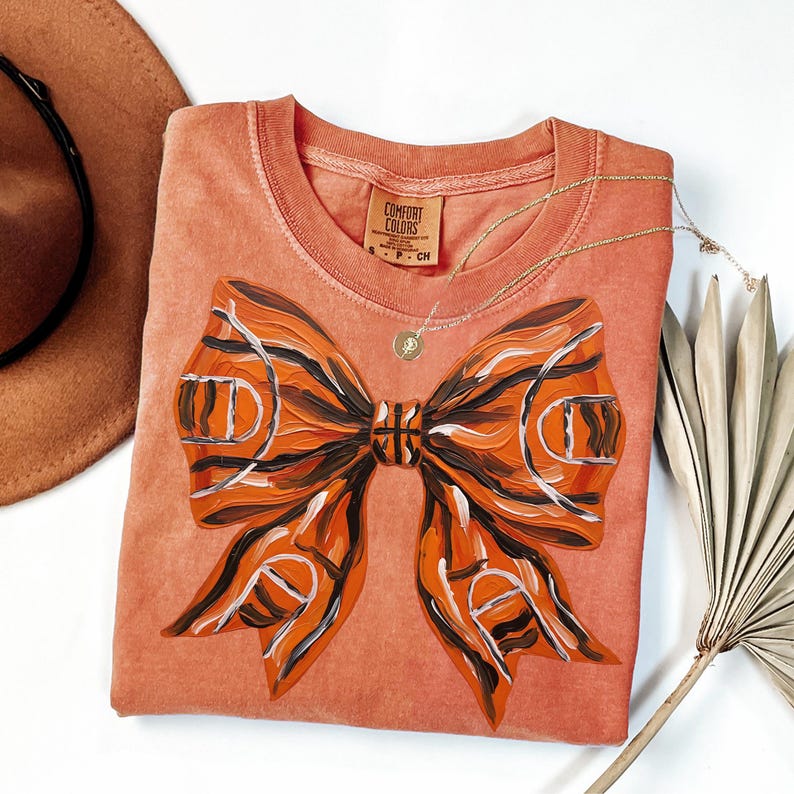 May include: A coral-colored Comfort Colors t-shirt featuring a large bow design. The bow is painted with orange, brown, and white stripes, resembling a basketball. A gold necklace with a small pendant is draped across the shirt.