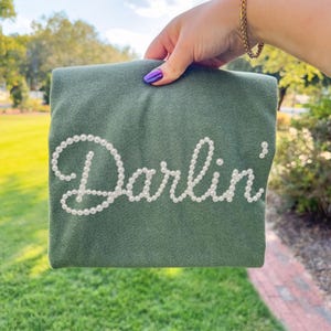 Pu&ograve; includere: Una felpa verde con la scritta "Darlin'" in perle bianche. La felpa &egrave; tenuta in mano con smalto viola, e si vede un braccialetto d'oro.