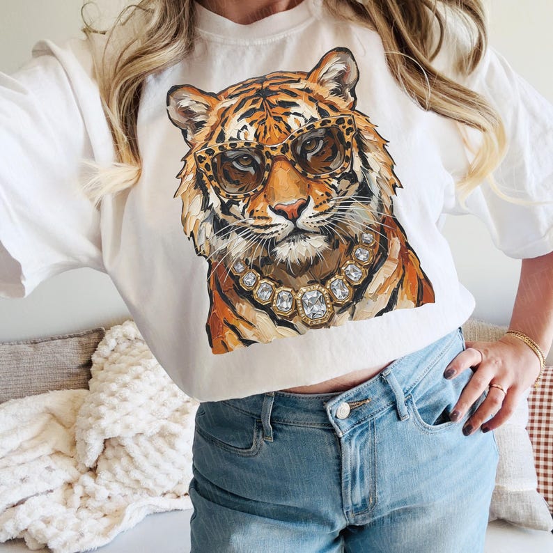 K&ouml;nnte beinhalten: Wei&szlig;es T-Shirt mit einer detaillierten Illustration eines Tigers mit Sonnenbrille und Diamantkette. Der Tiger hat orange und schwarze Streifen. Das Shirt wird mit einer blauen Jeans kombiniert.