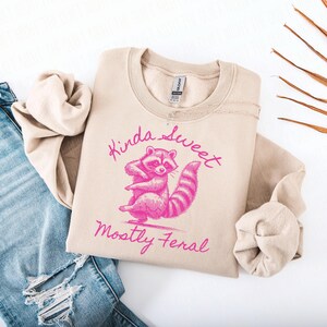 May include: Beige sweatshirt with a pink raccoon graphic and the text "Kinda Sweet Mostly Feral." The raccoon is standing with its arms crossed. The sweatshirt is folded next to a pair of blue jeans.
