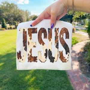 K&ouml;nnte beinhalten: Ein wei&szlig;es T-Shirt mit dem Wort "JESUS" im Kuhfellmuster. Die Buchstaben sind braun, schwarz und cremefarben und erzeugen ein rustikales und einzigartiges Design. Das Shirt wird vor einem Hintergrund aus gr&uuml;nem Gras und blauem Himmel gehalten.