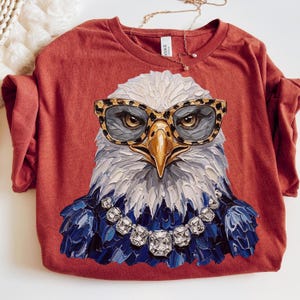 May include: A rust-colored t-shirt featuring a graphic of a bald eagle wearing leopard-print glasses and a silver-toned rhinestone necklace. The eagle is rendered in a thick, textured paint style with white, gray, and dark blue plumage details.