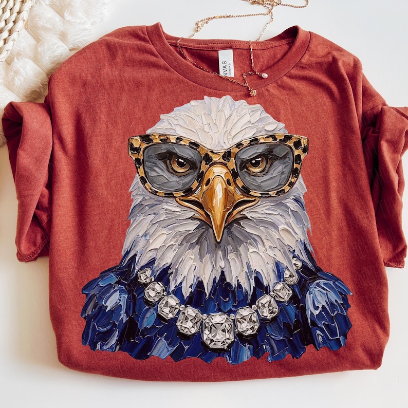May include: A rust-colored t-shirt featuring a graphic of a bald eagle wearing leopard-print glasses and a silver-toned rhinestone necklace. The eagle is rendered in a thick, textured paint style with white, gray, and dark blue plumage details.