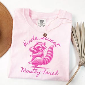 May include: A light pink t-shirt with a raccoon graphic and the text "Kinda Sweet, Mostly Feral" in pink script. The shirt has a crew neck and short sleeves. A gold necklace with a pendant is draped over the shirt.