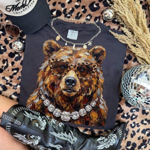 May include: A black t-shirt featuring a detailed illustration of a bear wearing leopard print glasses and a diamond necklace. The bear is brown and tan with a black nose. Accessories include a black hat, sunglasses, and a gold necklace.