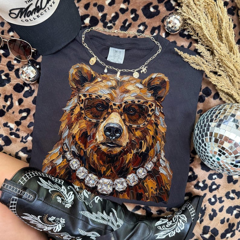 May include: A black t-shirt featuring a detailed illustration of a bear wearing leopard print glasses and a diamond necklace. The bear is brown and tan with a black nose. Accessories include a black hat, sunglasses, and a gold necklace.
