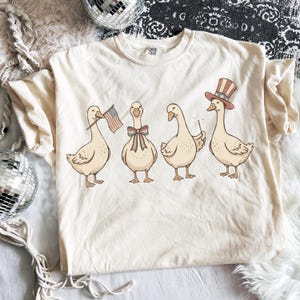 May include: Cream-colored t-shirt featuring a whimsical illustration of four geese. Each goose is adorned with patriotic accessories: an American flag, a bow, a sparkler, and a Uncle Sam hat. The shirt has a Comfort Colors tag.