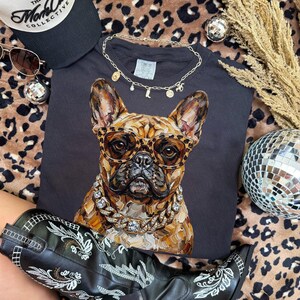 May include: Black t-shirt featuring a detailed illustration of a French Bulldog wearing leopard print glasses and a diamond chain. The shirt is accessorized with a gold necklace, sunglasses, and a black cap. A pair of black cowboy boots with white embroidery are also visible.
