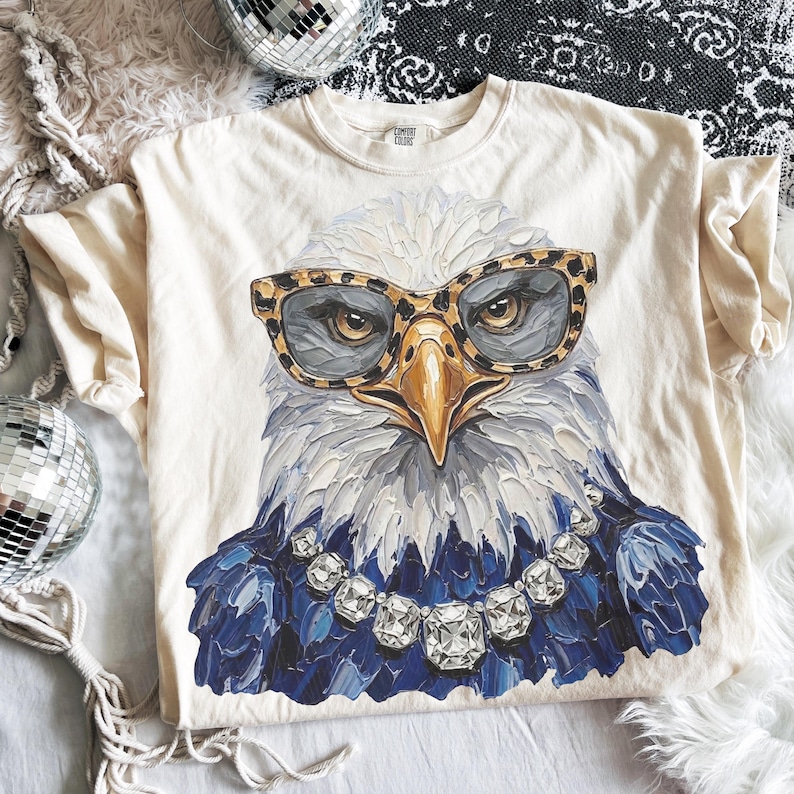 May include: A cream-colored t-shirt featuring a graphic of a bald eagle wearing leopard print glasses and a large, multi-faceted clear crystal necklace. The eagle has white head feathers and dark blue chest feathers, rendered in a textured, painterly style.