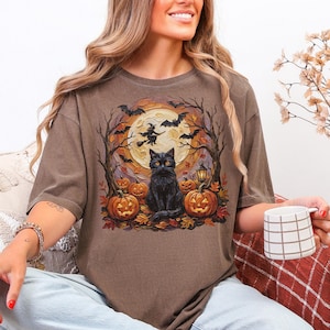 May include: A brown t-shirt featuring a graphic print of a black cat sitting in front of a large yellow moon. The design includes silhouettes of flying bats, a witch on a broomstick, bare trees, a lantern, and several orange jack-o'-lanterns.